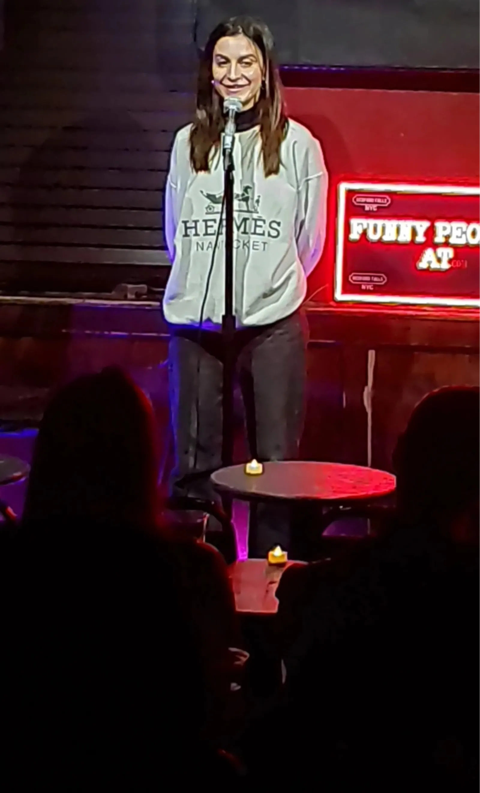 Comedian at the mic beside the red Funny People At neon sign at Upper East Side Comedy Club