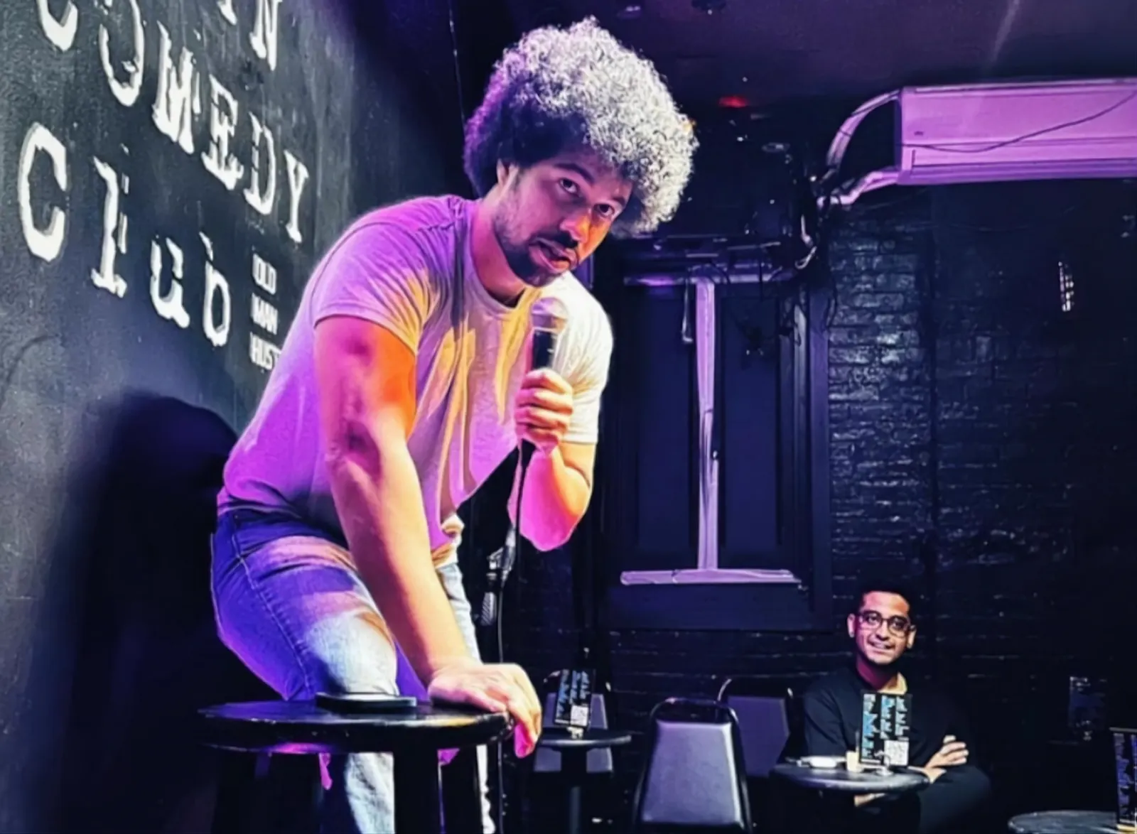 Comedian perched on a stool performing at Old Man Hustle BKLYN Comedy Club