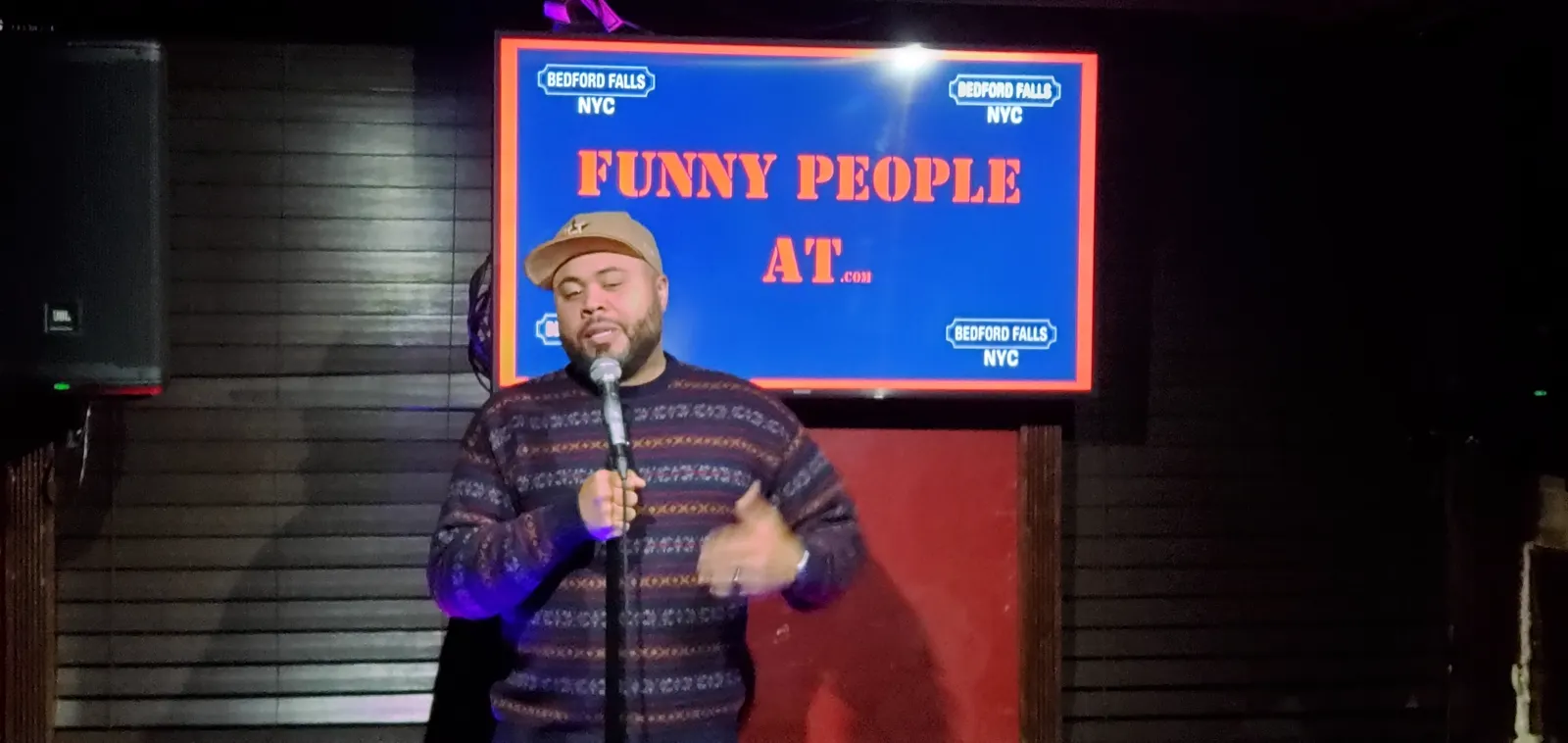 Comic in a striped sweater performing under the Funny People At screen at Upper East Side Comedy Club