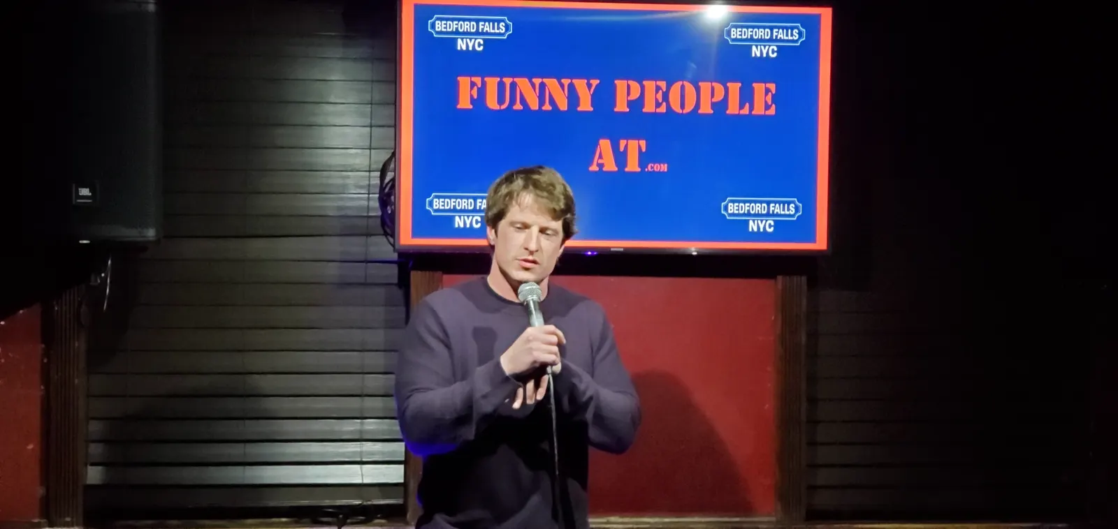 Stand-up comic on stage with the Funny People At screen behind him at Upper East Side Comedy Club